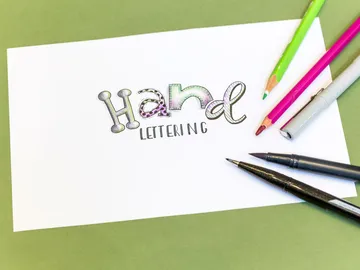 Buntstifte und Marker umgeben das Wort „Hand Lettering“.
